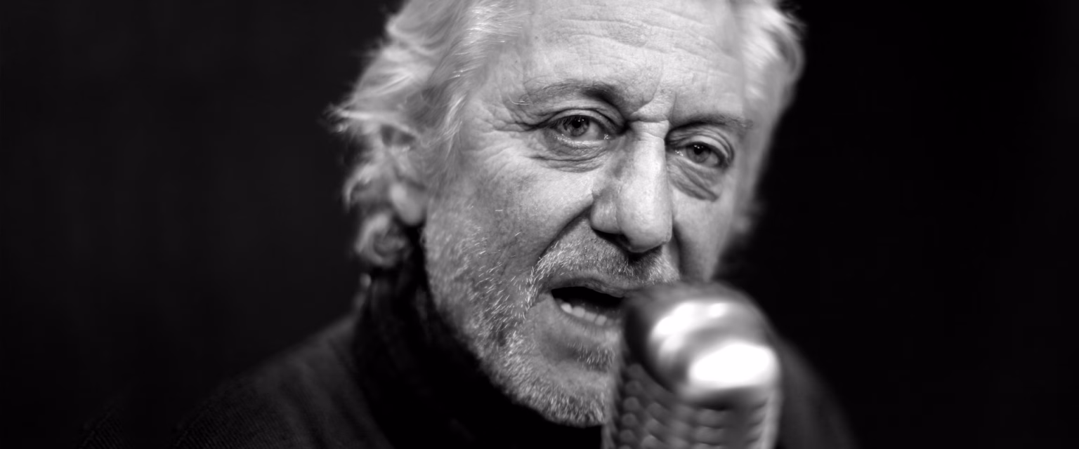 Portrait noir et blanc de Patrick Poivey, comédien voix off Sonacom, en col roulé, le micro vintage à la main