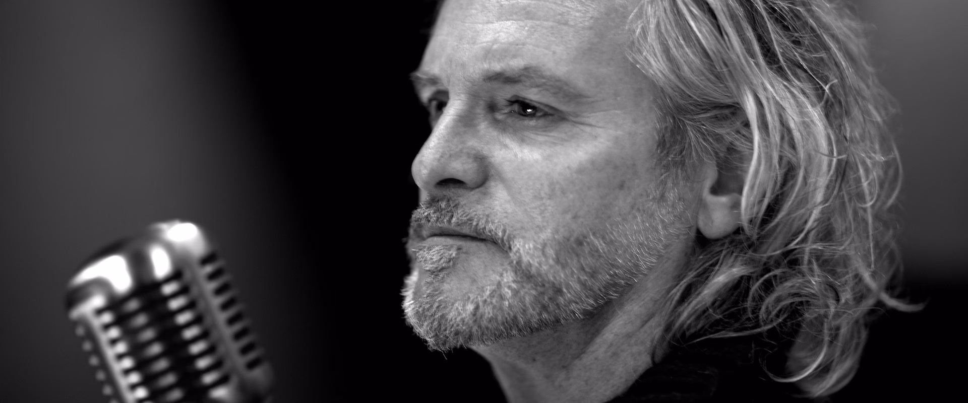 Portrait noir et blanc de Pierre-Alain de Garrigues, comédien voix off Sonacom, de profil, cheveux longs, au micro vintage